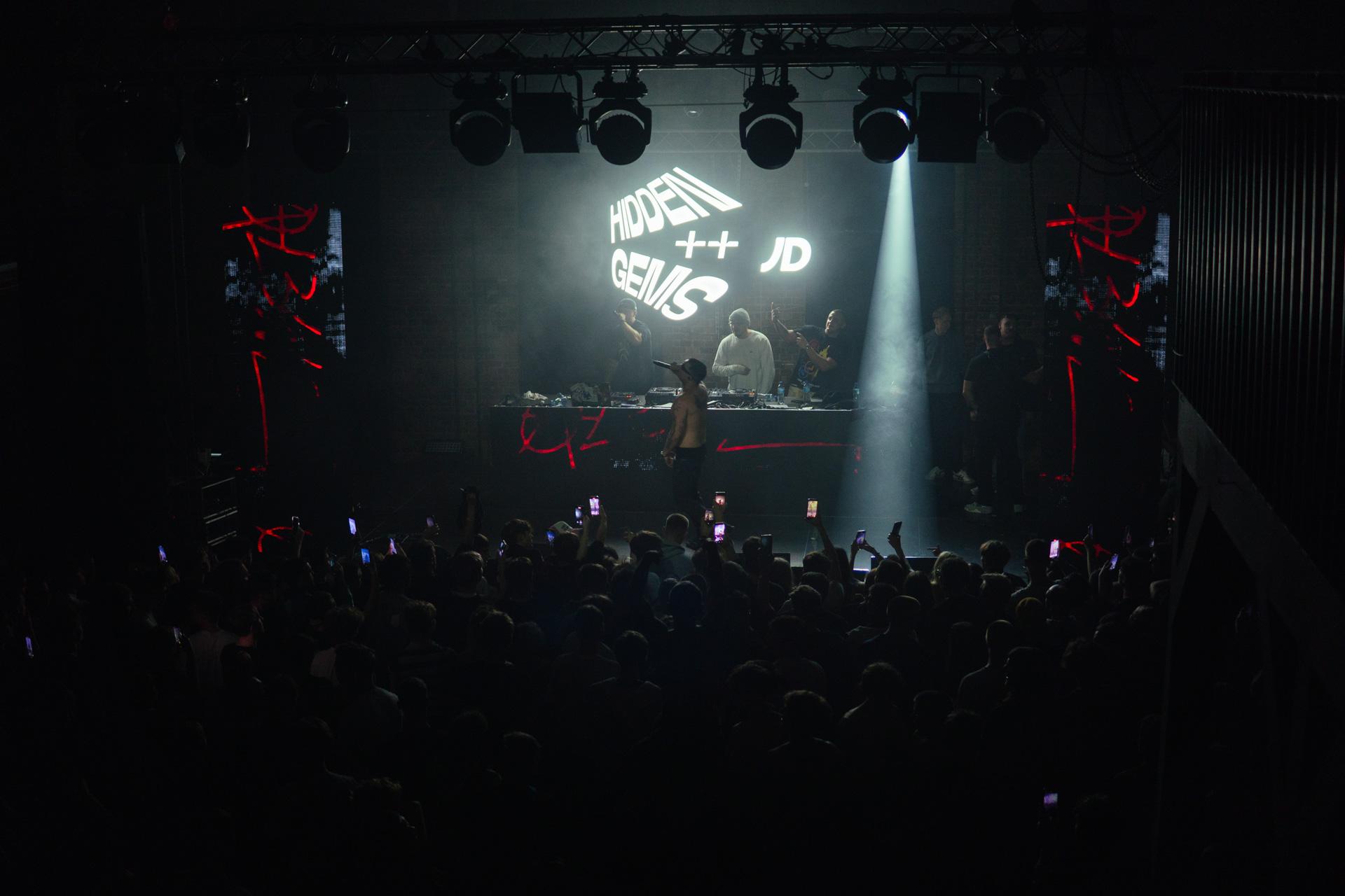 A wide, high-angle view of the dark concert venue, showing performers silhouetted against a bright screen displaying the 'Hidden Gems' logo while the audience records the show on their phones.