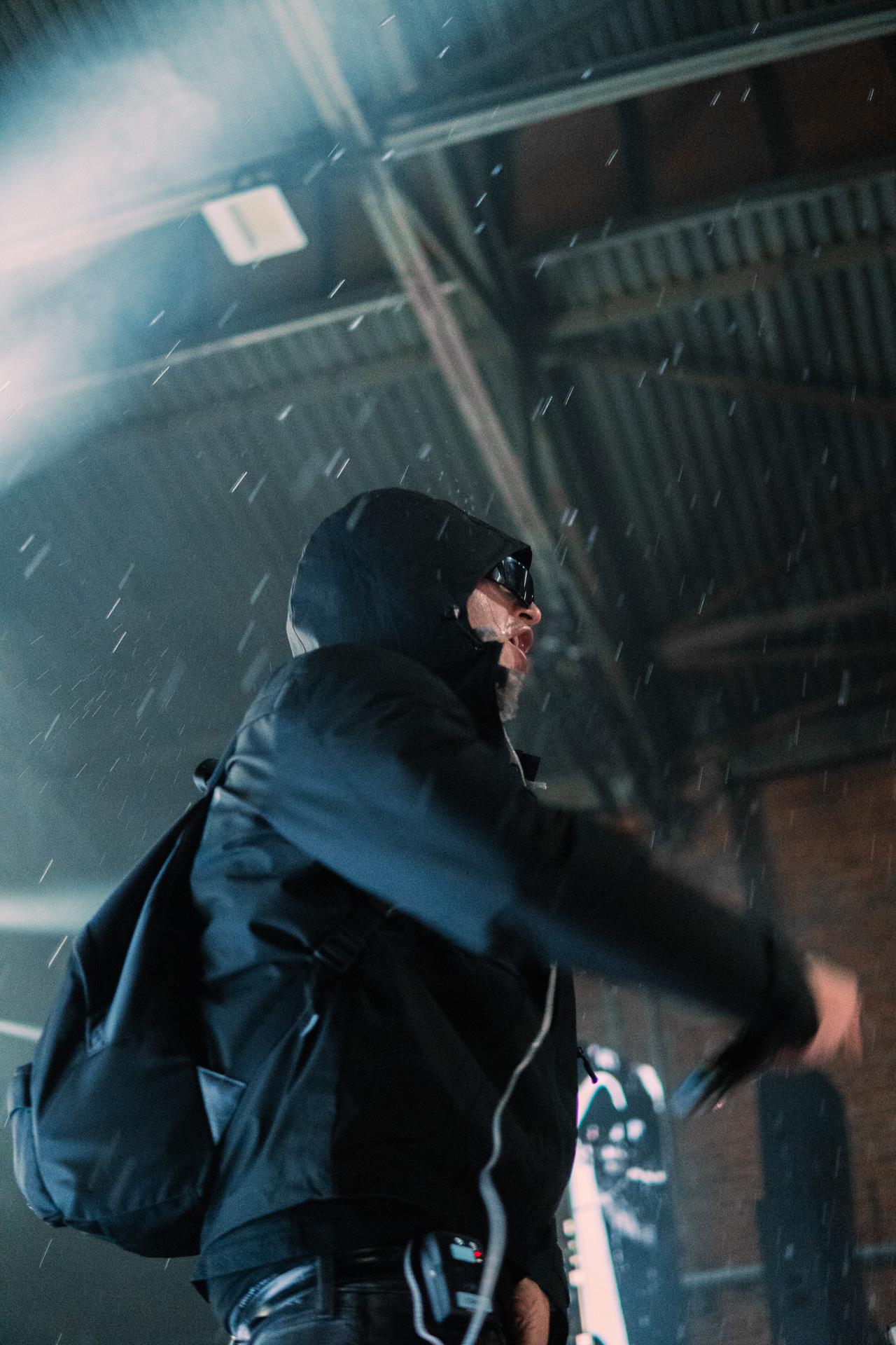A moody, low-angle profile shot of a performer wearing a black hoodie, sunglasses, and a backpack under an industrial ceiling with atmospheric water drops catching the light.