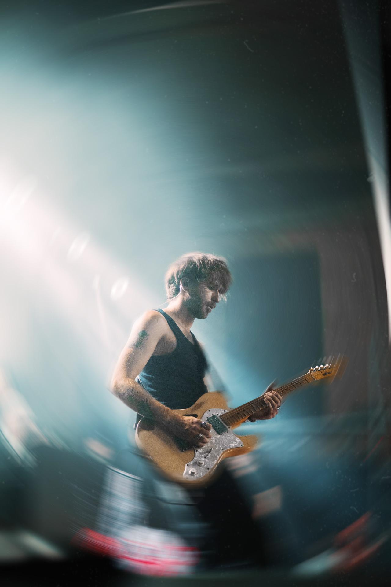 A concert photographer's shot of a musician playing an electric guitar on stage, illuminated by a dramatic, hazy spotlight with a creative radial blur effect.