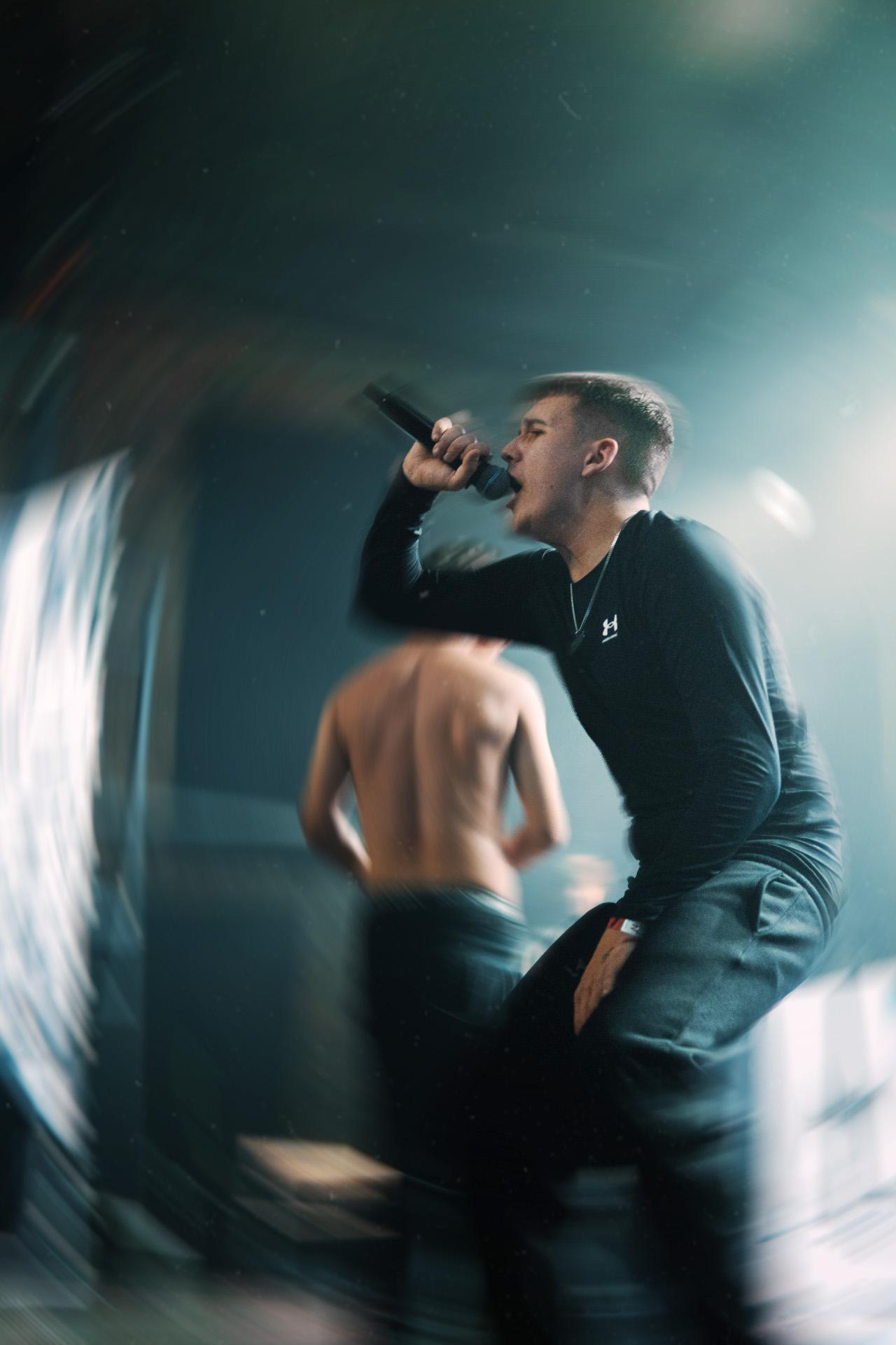 A dynamic, heavily motion-blurred shot of a vocalist in a black long-sleeve shirt passionately performing on stage, with another person visible in the soft-focus background.