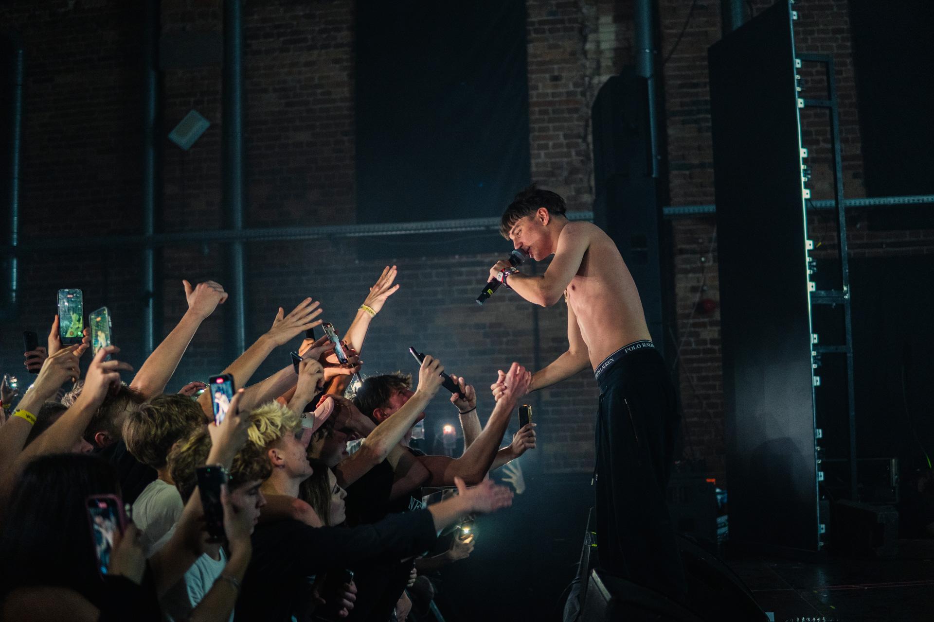 A shirtless performer on an elevated stage leans down to grasp the hands of excited fans in the front row, capturing a moment of strong artist-crowd connection against an industrial brick wall.