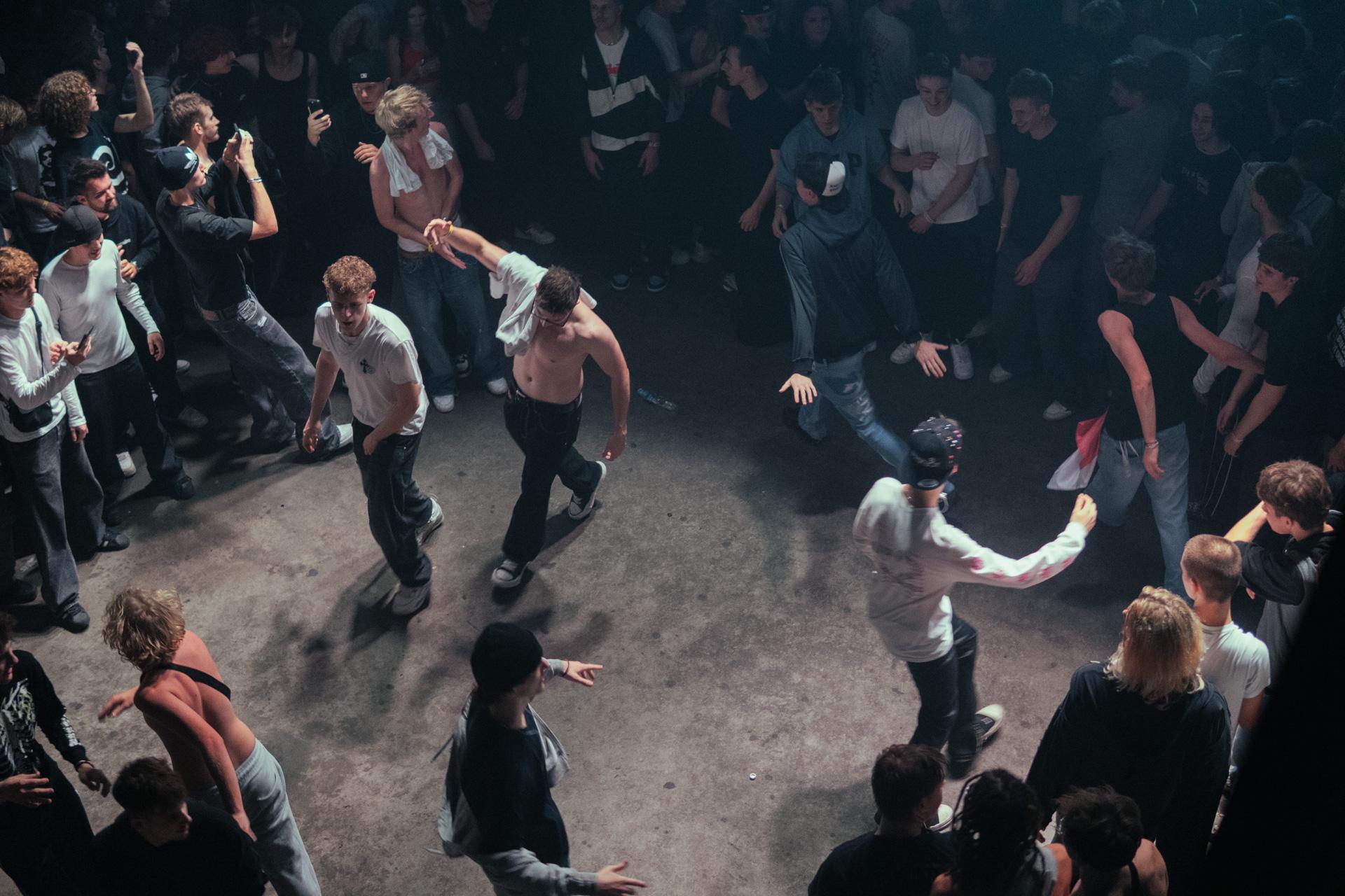 A high-angle, atmospheric view of an energetic mosh pit at a live music event, showing crowd members dancing intensely in a cleared circle.