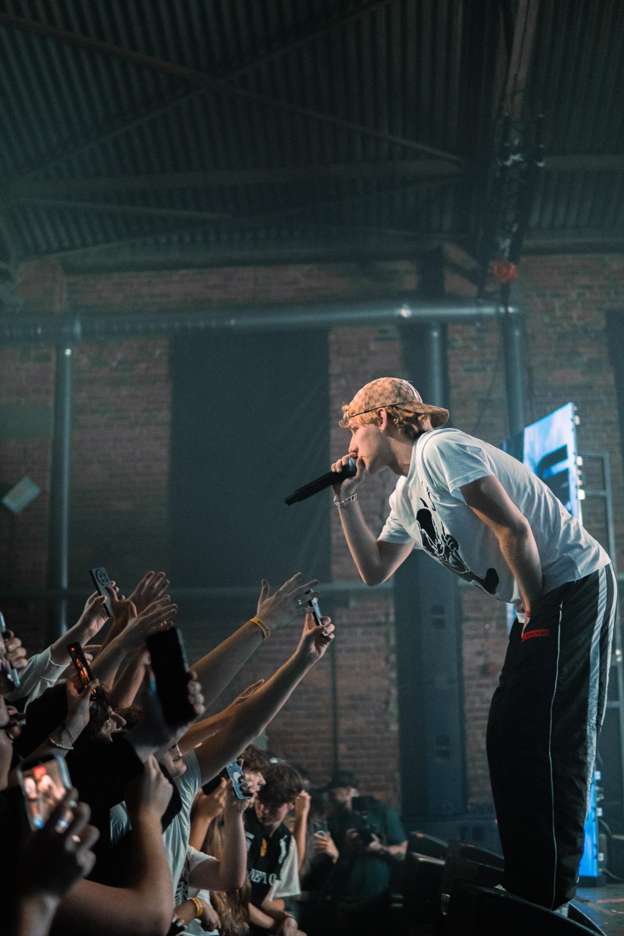 A side-profile shot of a performer wearing a cap and graphic t-shirt, leaning out over the edge of the stage to sing directly into a dense crowd of fans reaching out with their hands and cameras.