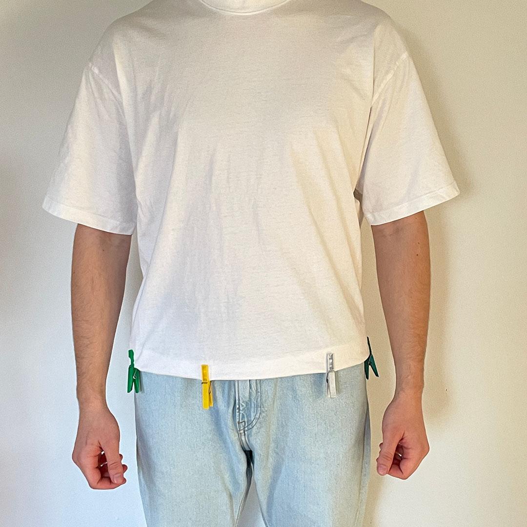 A person wearing a plain white oversized t-shirt, with clothespins attached to the bottom hem to demonstrate the garment's fit and proportions