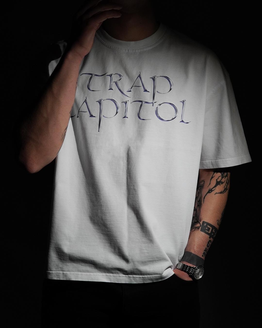 Model in a moody, dark studio setting wearing a white t-shirt showcasing the metallic 'TRAP CAPITOL' typography graphic on the chest