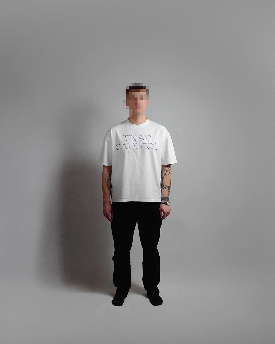 Front view of a tattooed model standing against a grey studio backdrop, wearing a white t-shirt with the 'TRAP CAPITOL' chest print